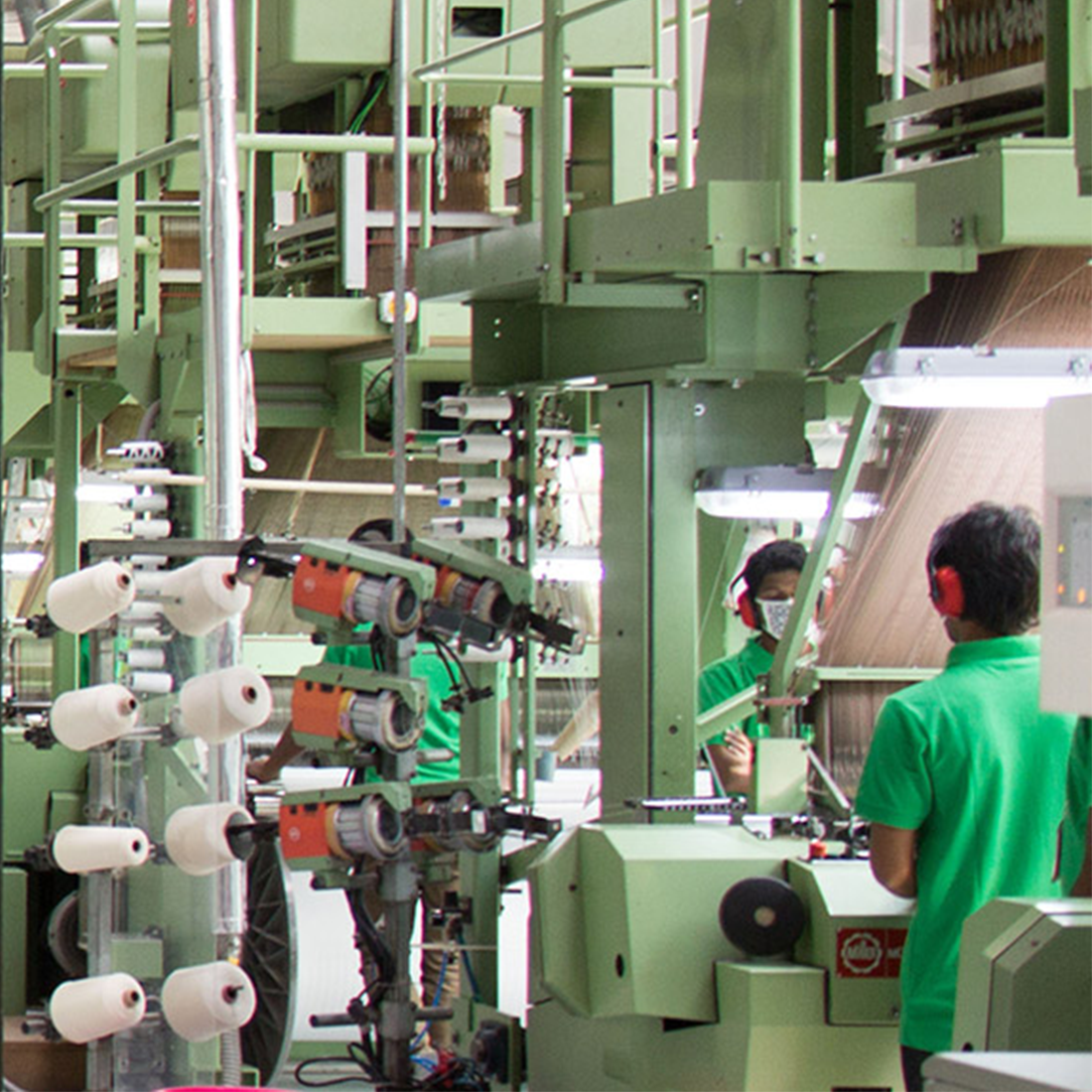 Man sieht eine Produktionshalle mit diversen Maschinen, wo Webetiketten gefertigt werden. An den Maschinen stehen Männer mit grünen Shirt und sie tragen alle einen roten Gehörschutz
